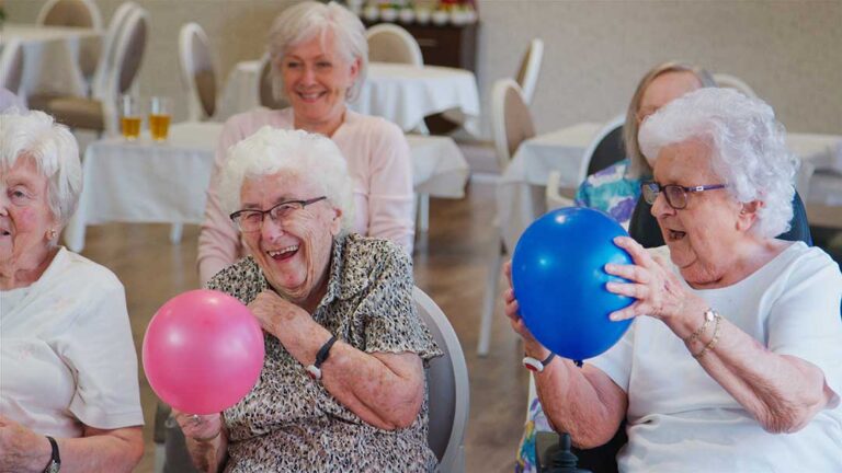 A group of older adults chatting at a table in a clean, well-lit common area with a caregiver nearby.